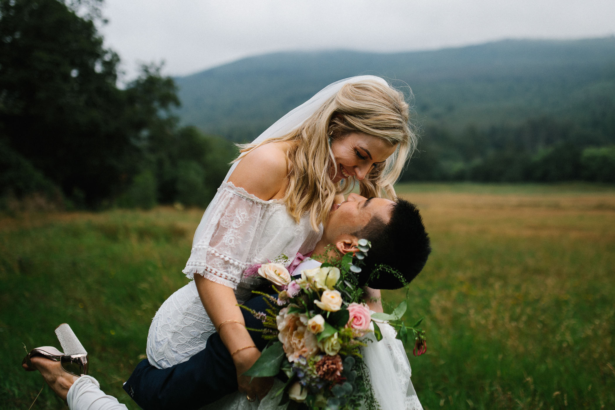 A bride is lifted up by her groom and they're about to kiss. She's holding a bouquet of ivory and pink flowers. By Luke Flint Photography